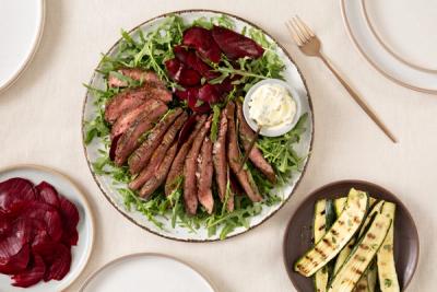 A main dish of sliced steak, rocket, beets, and a small bowl of sauce, served with sides of grilled zucchini and sliced beets.