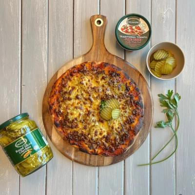 A cheeseburger pizza with ground meat, cheese, and pickles on a wooden board, accompanied by a jar of pickles, a bowl of pickles, and a container of pizza sauce. A cheeseburger pizza with ground meat, cheese, and pickles on a wooden board, accompanied by a jar of pickles, a bowl of pickles, and a container of pizza sauce.