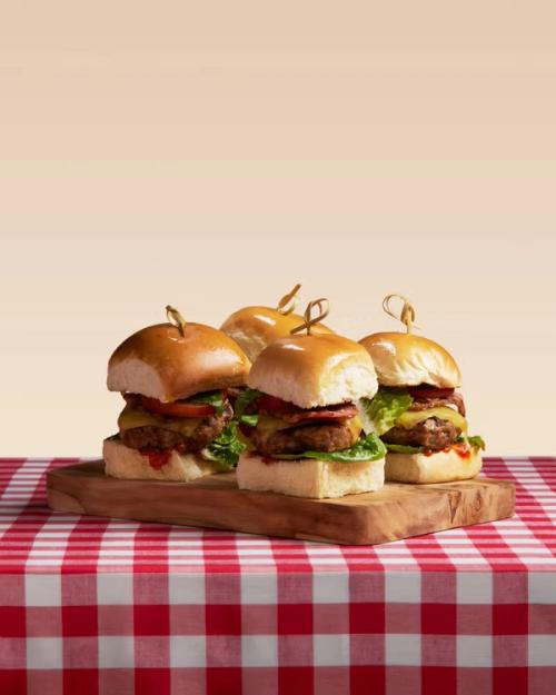 Four cheeseburger sliders on a wooden board on a red and white checkered tablecloth.