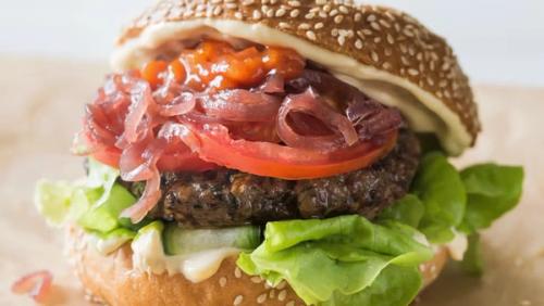 A gourmet burger on a sesame seed bun with a dark patty, lettuce, cucumber, tomato, pickled red onions, and a red relish. A gourmet burger on a sesame seed bun with a dark patty, lettuce, cucumber, tomato, pickled red onions, and a red relish.