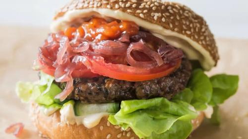 A gourmet burger on a sesame seed bun with a dark patty, lettuce, cucumber, tomato, pickled red onions, and a red relish.