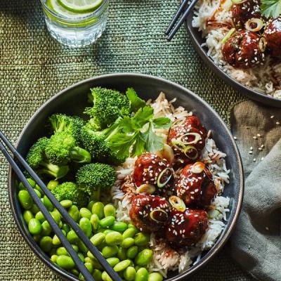 a bowl of rice, broccoli, edamame and meatballs with chopsticks on a table. a bowl of rice, broccoli, edamame and meatballs with chopsticks on a table.