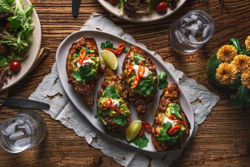 Four stuffed sweet potatoes topped with guacamole, sour cream, and chili, served on a wooden table with a side salad.