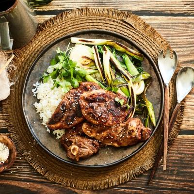 Grilled lamb chops with sesame seeds, white rice, charred green onions, and cilantro on a dark plate.