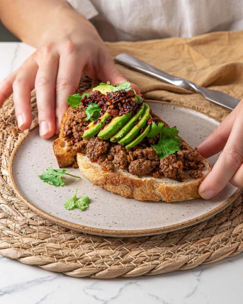 a person is holding a piece of toast with mince and avocado on a plate . a person is holding a piece of toast with mince and avocado on a plate .