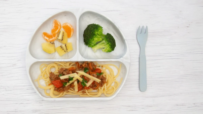 A child's divided plate with spaghetti bolognese, broccoli florets, and a fruit mix of orange segments and kiwi slices, next to a light blue fork.