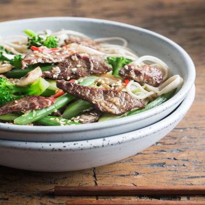a bowl of beef and broccoli with noodles and chopsticks on a wooden table .