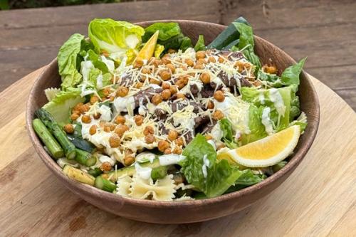 A large salad in a wooden bowl with lettuce, sliced steak, roasted chickpeas, shredded cheese, asparagus, bowtie pasta, and a lemon wedge.