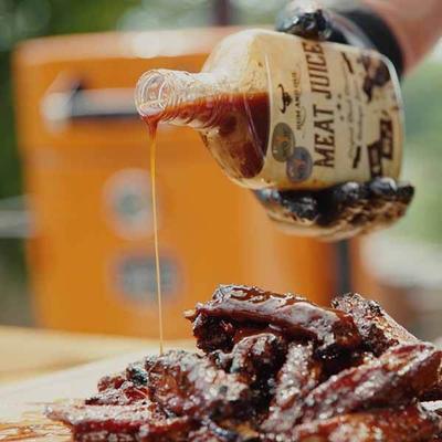 A hand in a black glove pours "MEAT JUICE" sauce from a bottle onto a pile of glistening barbecued lamb ribs. A hand in a black glove pours "MEAT JUICE" sauce from a bottle onto a pile of glistening barbecued lamb ribs.