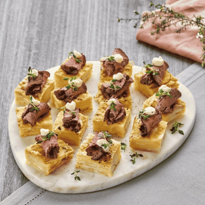 Roast beef and layered potato canapés garnished with cream and thyme on a white marble board.