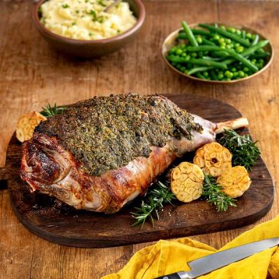 a large piece of lamb is sitting on top of a wooden cutting board with a bowls of beans and mashed potato on a table. 