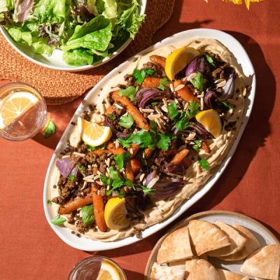 A platter of hummus topped with roasted carrots, red onions, spiced lamb, parsley, lemon wedges, and almonds, served with a green salad, pita bread, and lemon water.