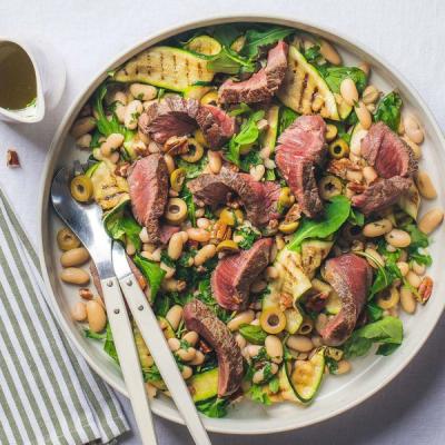 a white plate topped with a salad of beans, steak, zucchini and olives