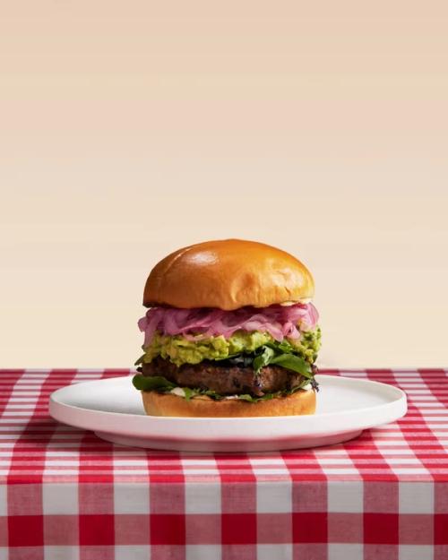A burger with a patty, greens, guacamole, and pickled red onions on a white plate on a red and white checkered tablecloth.