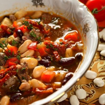 Hearty bean and meat soup with carrots, sun-dried tomatoes, and thyme in an ornate white bowl.