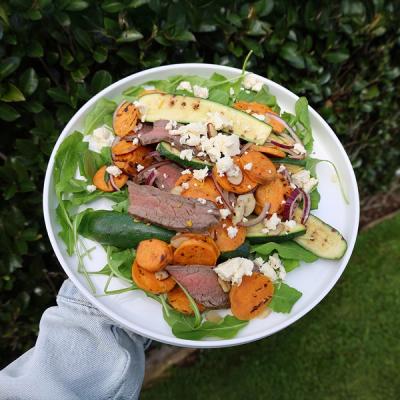 a person is holding a white plate with a salad of grilled steak, kumara and courgette.