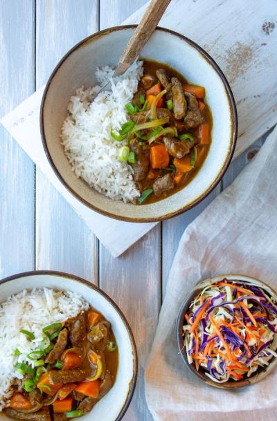 two bowls of beef curry with rice and a bowl of coleslaw on a table . two bowls of beef curry with rice and a bowl of coleslaw on a table .