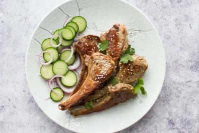 Grilled lamb chops with sesame seeds and cilantro, served with sliced cucumber and red onion on a white plate.