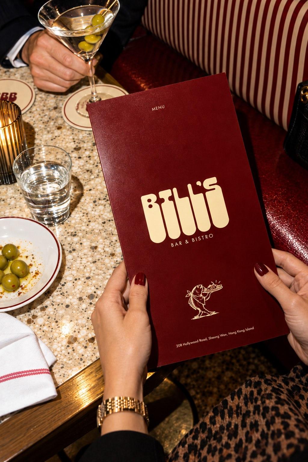 A Bill’s menu held open over a terrazzo table with olives, water and a martini