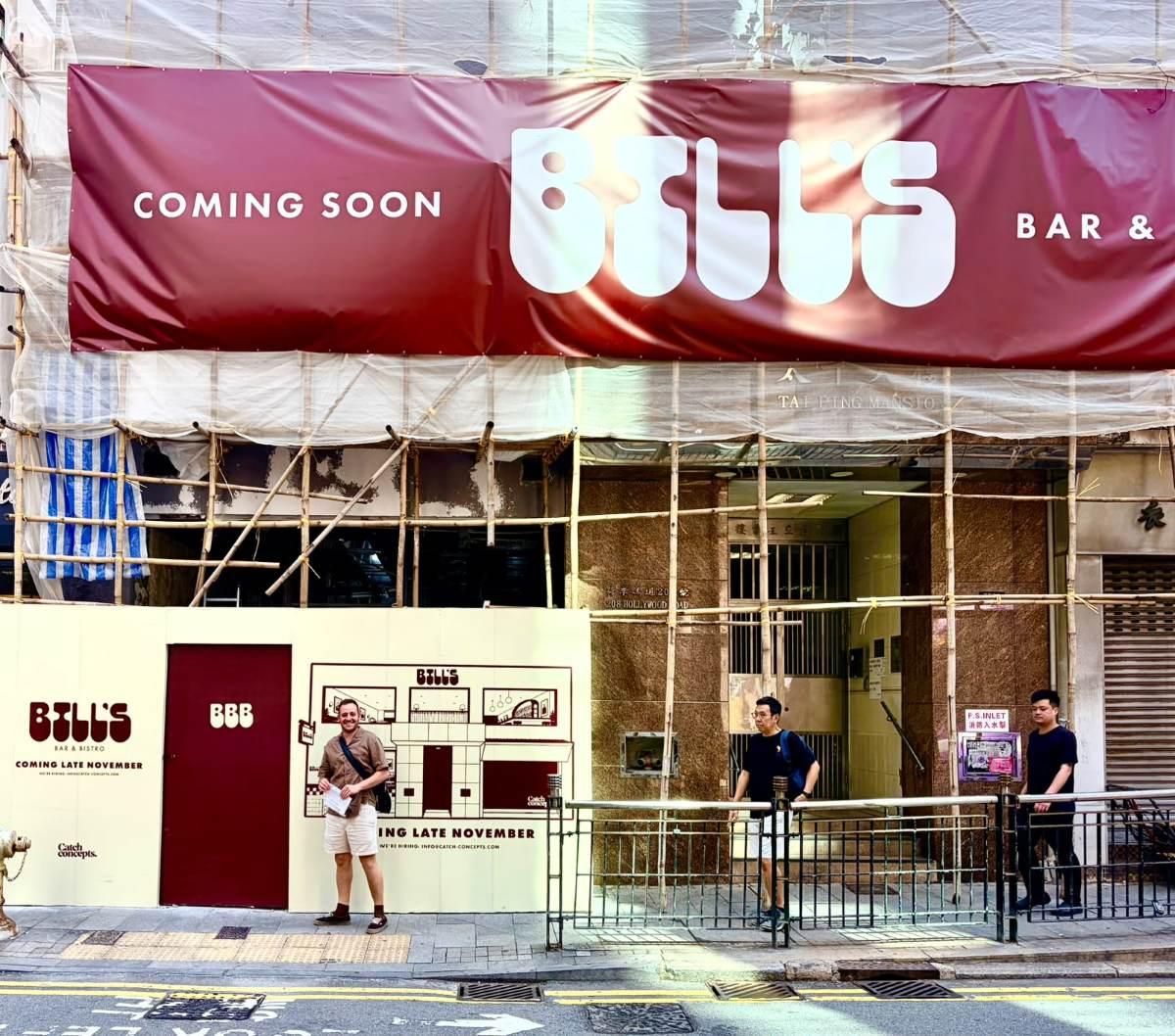 The red Bill’s Coming soon banner over bamboo scaffolding on Hollywood Road, with the Bill’s hoarding at street level and a man standing out front