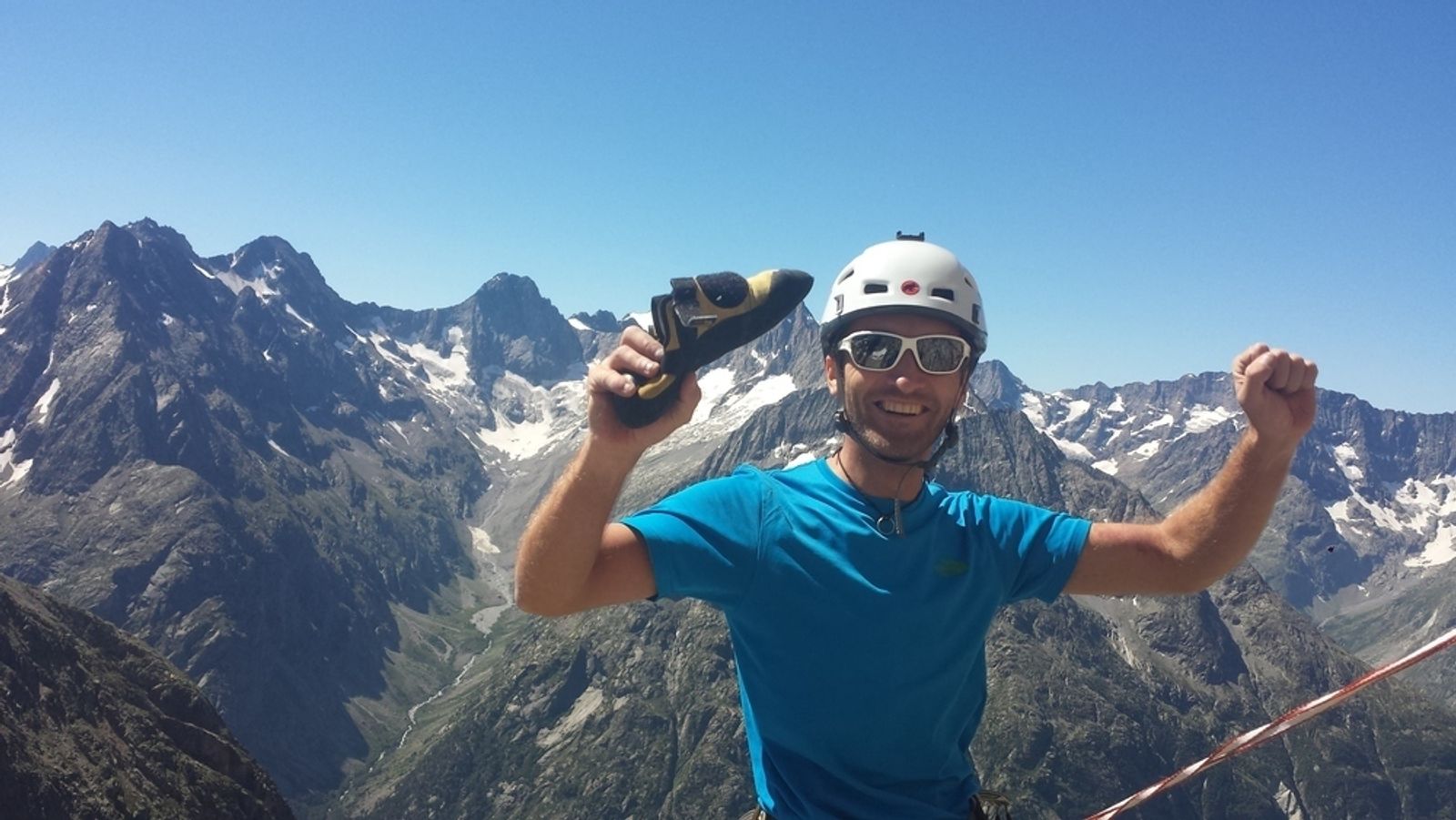 Arnaud sourit en brandissant ses chaussons après l’escalade, avec les montagnes du Soreiller derrière lui.