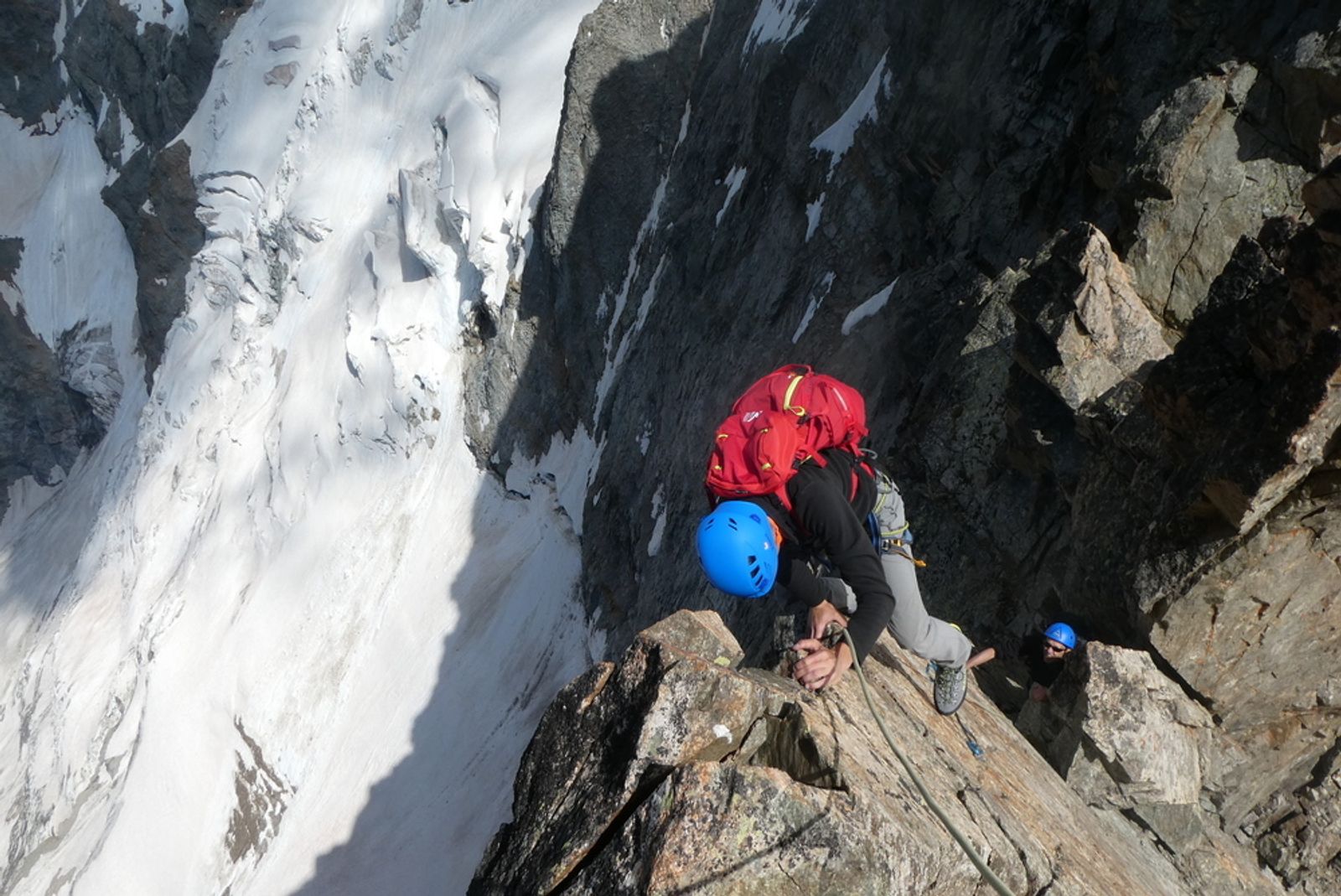 Alpiniste au sac rouge escaladant une arête rocheuse des Trifides au-dessus d’un versant raide et enneigé.