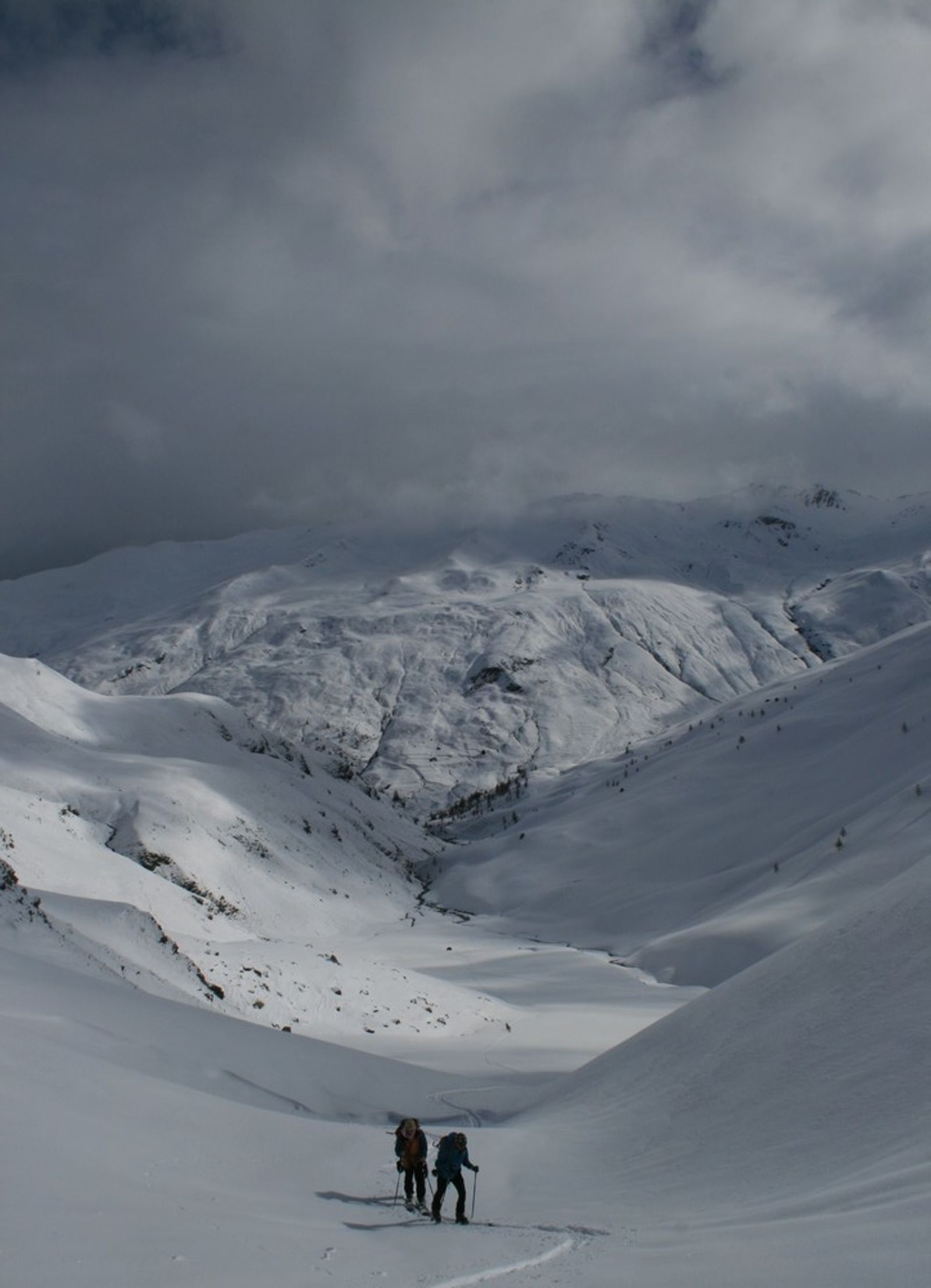 Two skiers climbing toward the Cime du Grand Vallon beneath dark clouds.