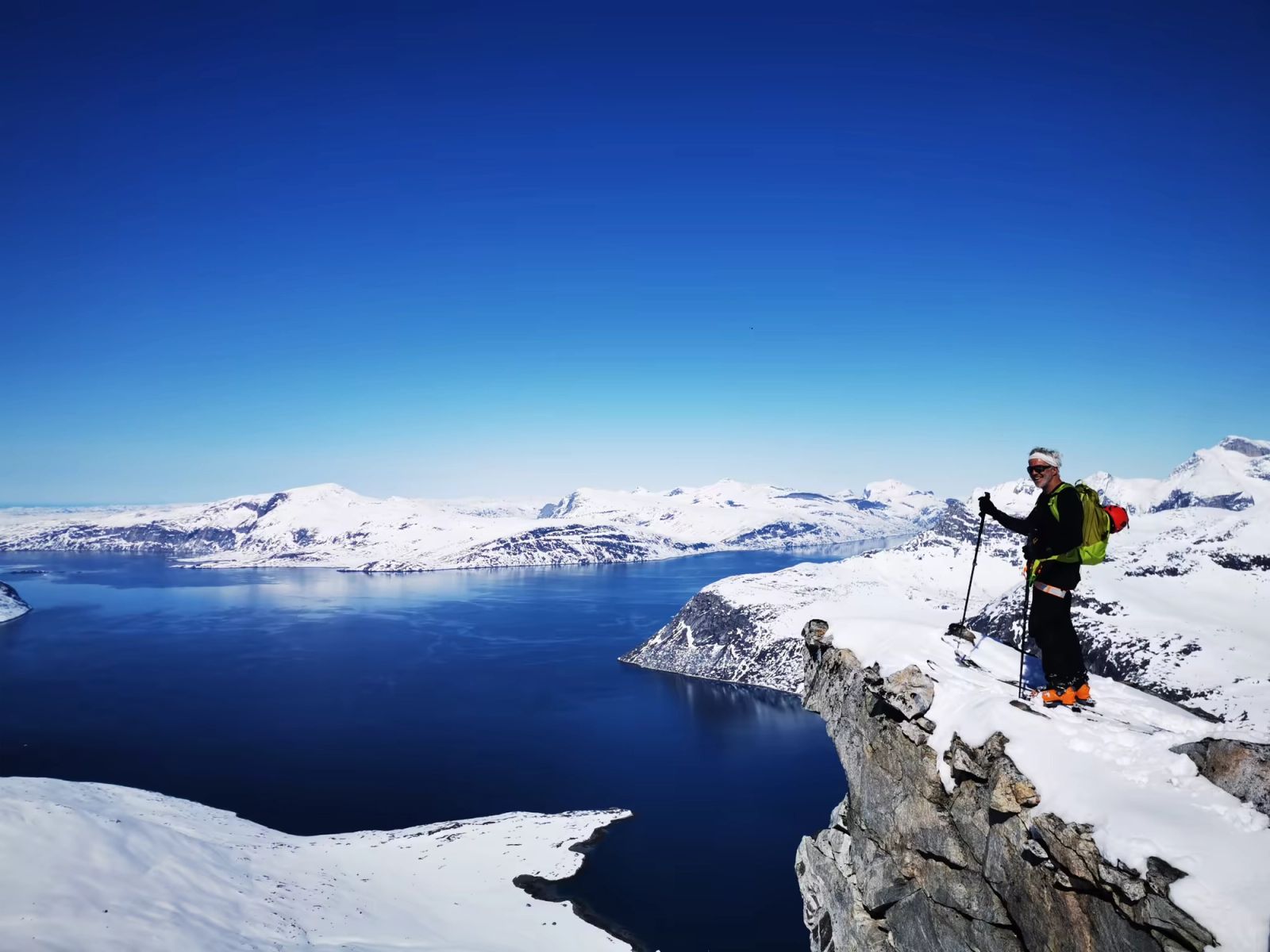 Skieur sur un belvédère rocheux dominant un fjord et ses montagnes enneigées au Groenland