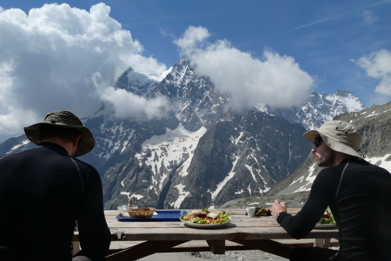 Nico et Tom prennent un repas sur la terrasse d’un refuge, face aux sommets et aux glaciers des Écrins.