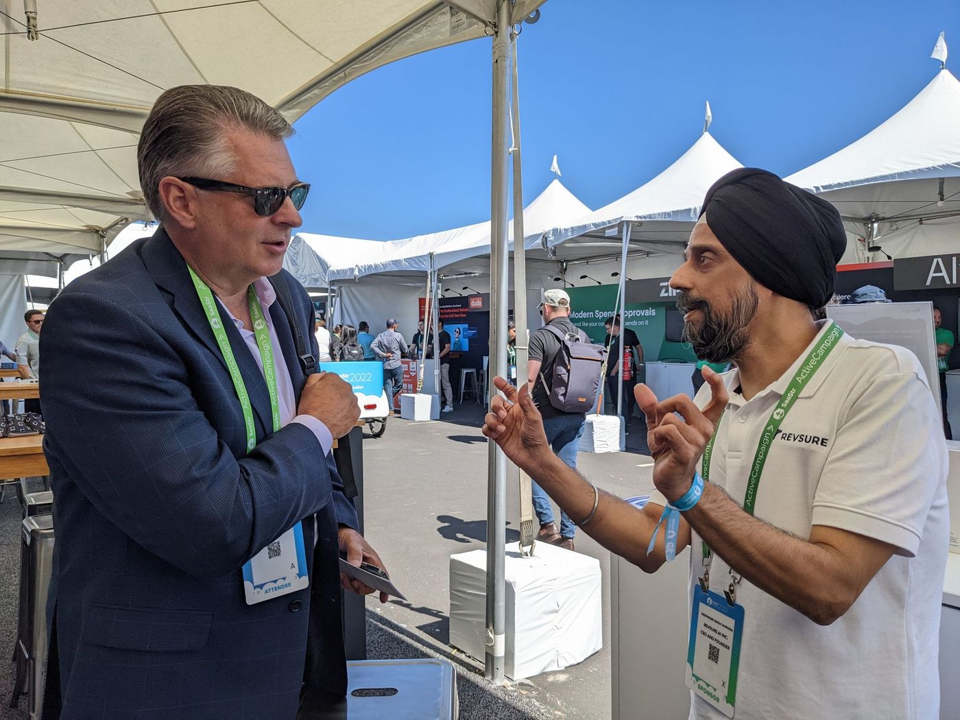 Two men in business casual attire with lanyards talking outdoors under a tent.