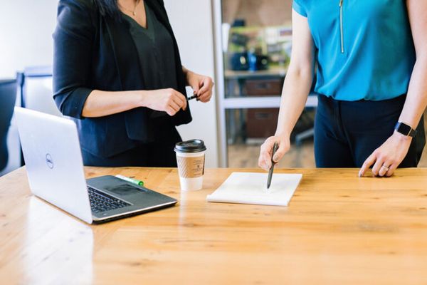 Two people stood at a desk with a laptop, cup of coffee and a sheet of paper being pointed at