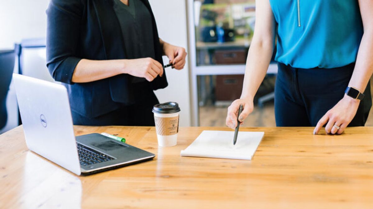 Two people stood at a desk with a laptop, cup of coffee and a sheet of paper being pointed at