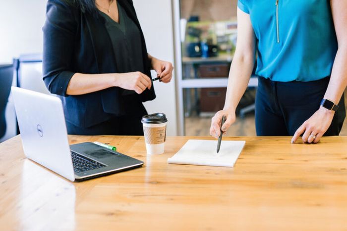 Two people stood at a desk with a laptop, cup of coffee and a sheet of paper being pointed at