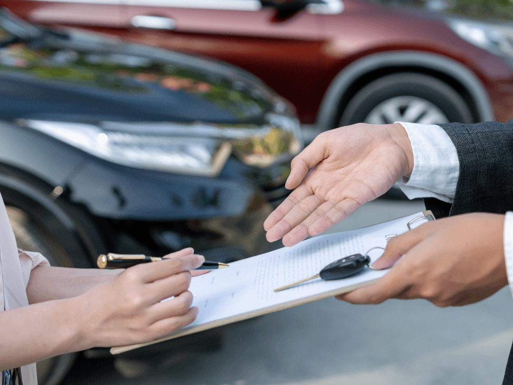 Negociação de veículo com documentos, caneta e chave sobre uma prancheta diante de carros estacionados.