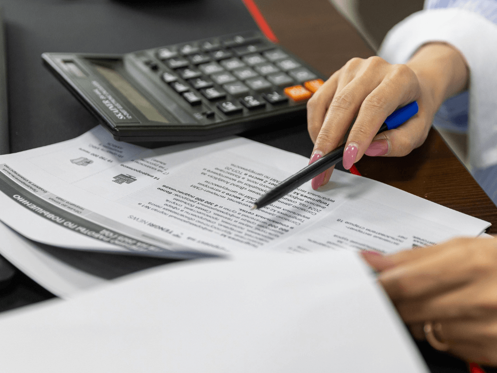 Pessoa utiliza uma caneta para analisar documentos impressos ao lado de uma calculadora durante uma atividade de conferência financeira.