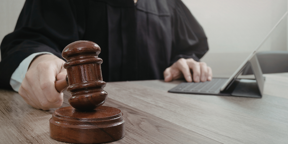 A imagem mostra um juiz ou autoridade judicial utilizando um martelo de tribunal (malhete), com um tablet ao fundo. O cenário transmite contexto jurídico, decisão judicial e processos legais.