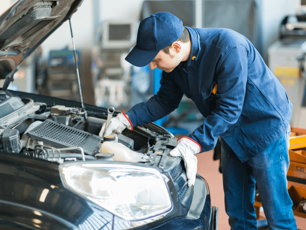 Mecânico com uniforme e boné azuis utiliza uma ferramenta para verificar componentes do motor de um carro com o capô aberto.