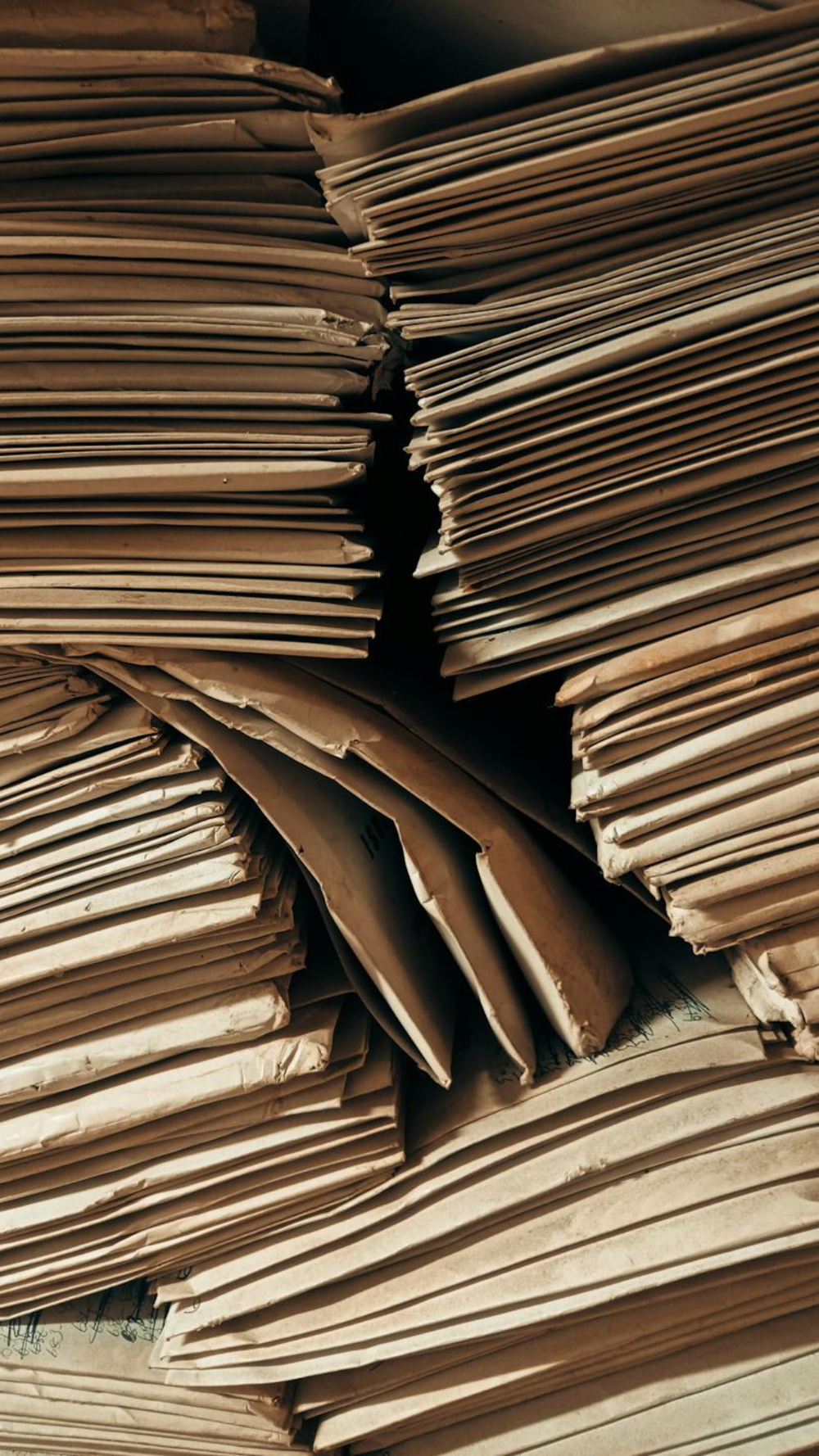 Pilhas densas de envelopes e pastas de papel pardo antigas, com bordas desgastadas e sinais de manuseio, organizadas de forma irregular em um arquivo