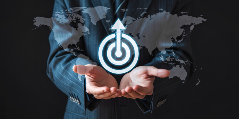 Mãos segurando símbolo de alvo com seta e mapa ao fundo, representando alcance de objetivos, estratégia global e precisão em metas no ambiente digital e de negócios