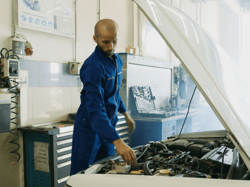 Mecânico de uniforme azul verifica o compartimento do motor de um carro em uma oficina durante uma inspeção automotiva.