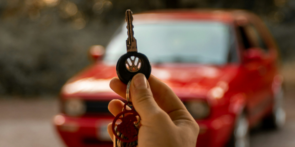Mão em primeiro plano segurando um molho de chaves com o logotipo da Volkswagen em destaque, com um carro Volkswagen Golf MK2 vermelho desfocado ao fundo em uma estrada arborizada