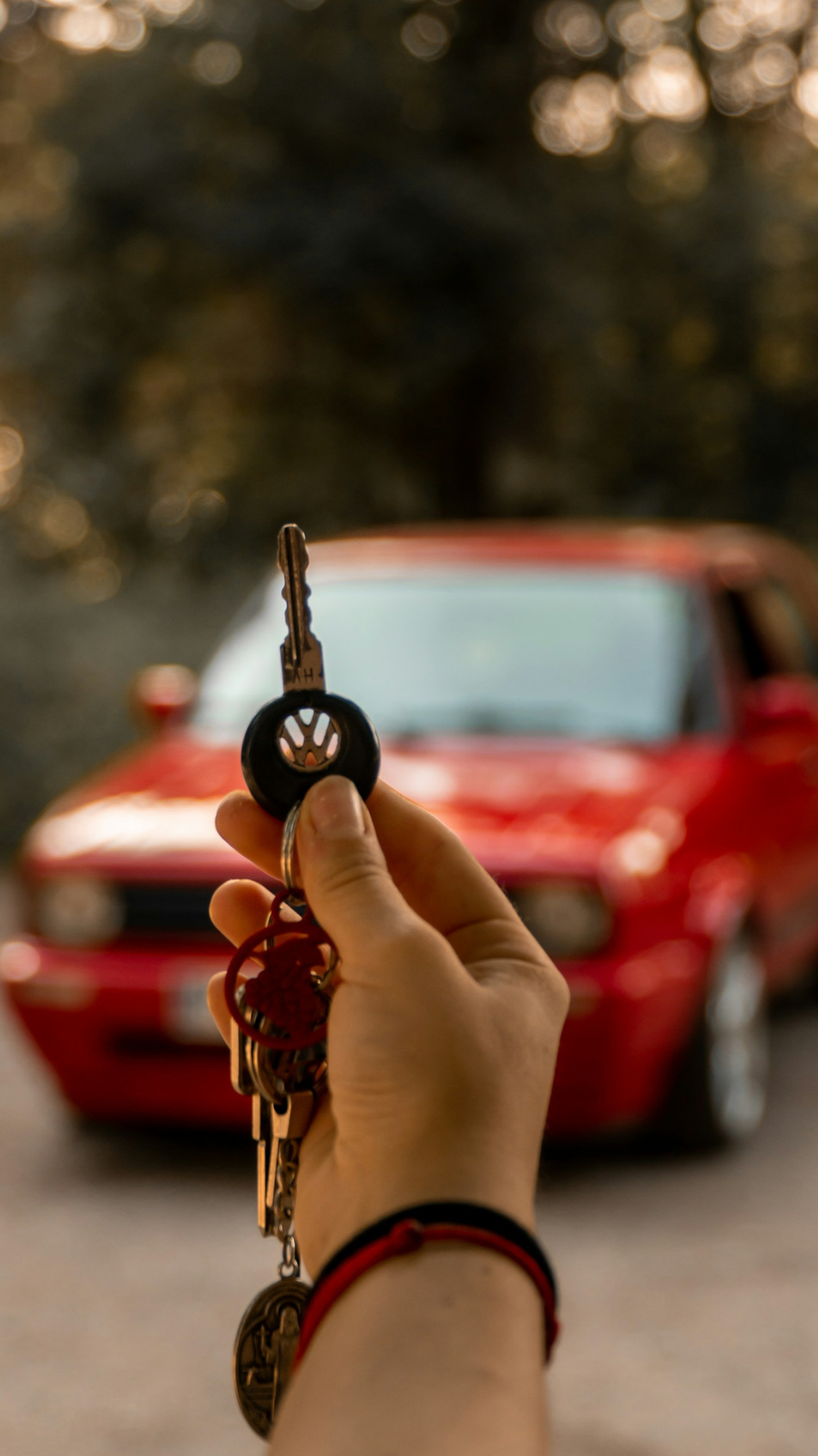 Mão em primeiro plano segurando um molho de chaves com o logotipo da Volkswagen em destaque, com um carro Volkswagen Golf MK2 vermelho desfocado ao fundo em uma estrada arborizada