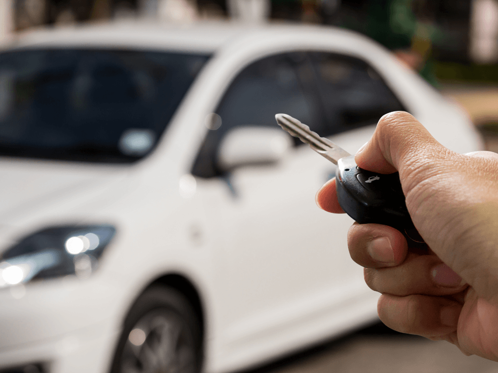 Pessoa segurando a chave de um carro diante de um automóvel branco, situação relacionada à compra de veículo usado. Tudo para explicar o que é histórico veicular.