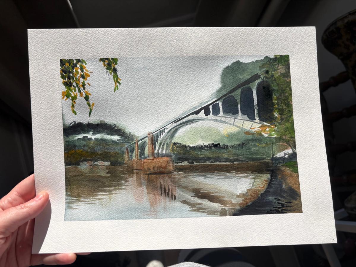 The 40th Street Bridge watercolor held in hand before framing, a white arch bridge over the Allegheny River with autumn leaves hanging in the corner