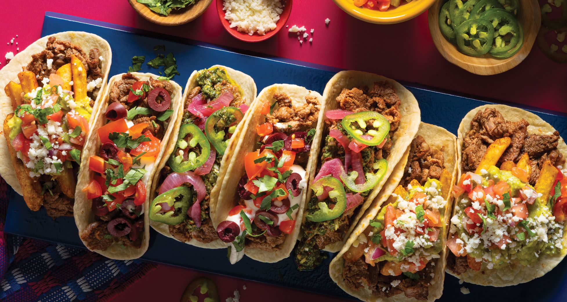 A row of seven soft-shell tacos loaded with seasoned meat, french fries, pico de gallo, cotija cheese, sliced jalapeños, olives, and pickled red onions on a blue platter.
