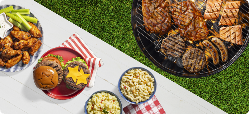 A grill with burger, pork steaks and a table with wings, a burger, and mac and cheese salad.