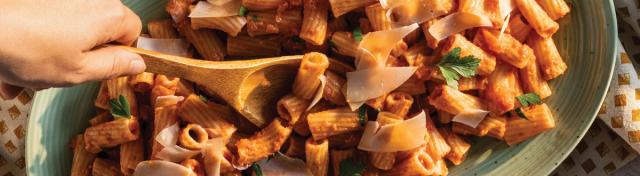 A large oval green platter filled with rigatoni pasta tossed in red sauce, garnished with shaved parmesan and fresh parsley, with a wooden spoon serving a portion.