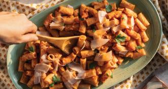 A large oval green platter filled with rigatoni pasta tossed in red sauce, garnished with shaved parmesan and fresh parsley, with a wooden spoon serving a portion.