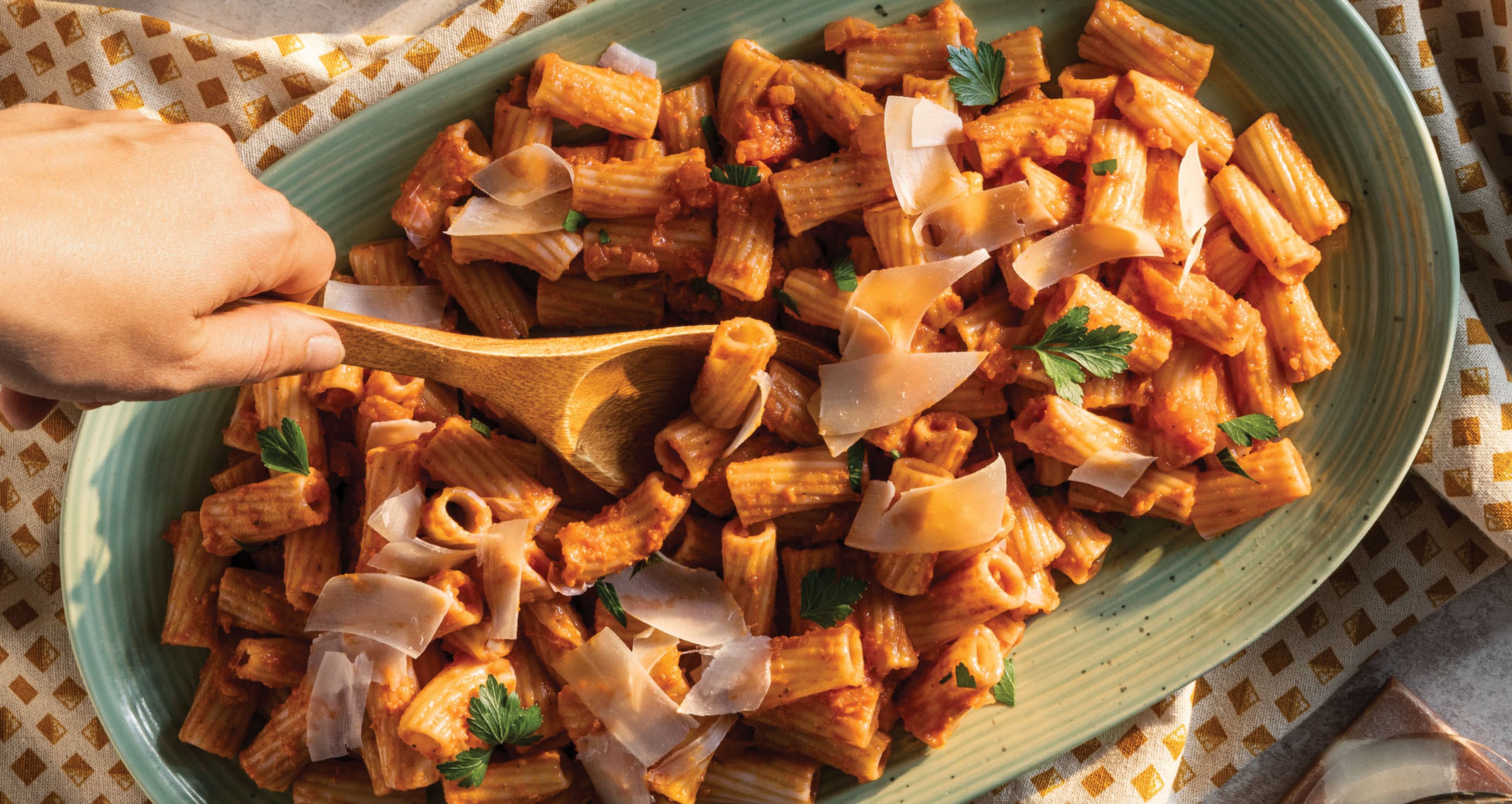 A large oval green platter filled with rigatoni pasta tossed in red sauce, garnished with shaved parmesan and fresh parsley, with a wooden spoon serving a portion.