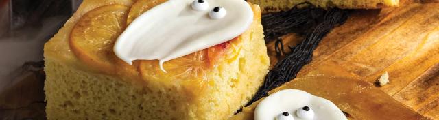 Square slices of citrus upside-down cake decorated with white icing ghosts with candy eyes for Halloween, served on a wooden board.